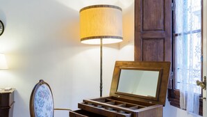 Una habitación con un escritorio de madera, una silla con estampado floral, una lámpara y una ventana con cortinas de encaje.