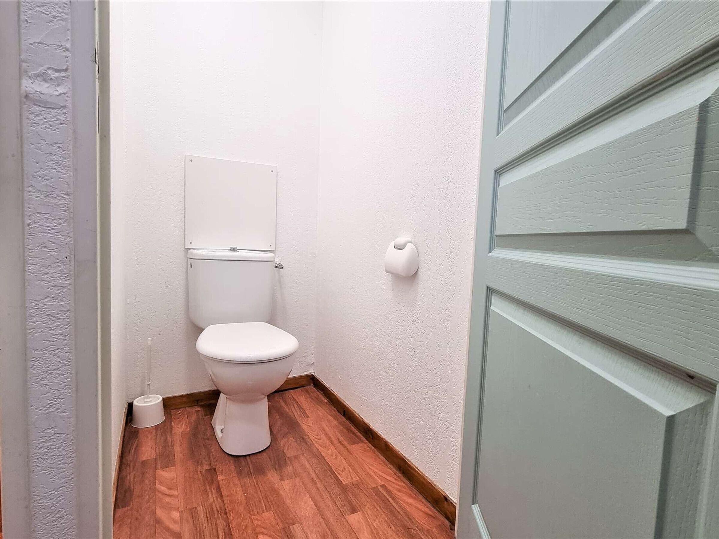 Een kleine, schone badkamer met een wit toilet, houten vloeren en een effen witte wand.