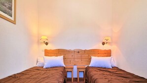 A room with two single beds, each with a wooden headboard, white pillows, and a brown bedspread. There are two wall-mounted lamps on either side of the headboard.