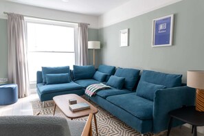 Un salón con un sofá azul, una mesa de centro, una lámpara y un cuadro en la pared.