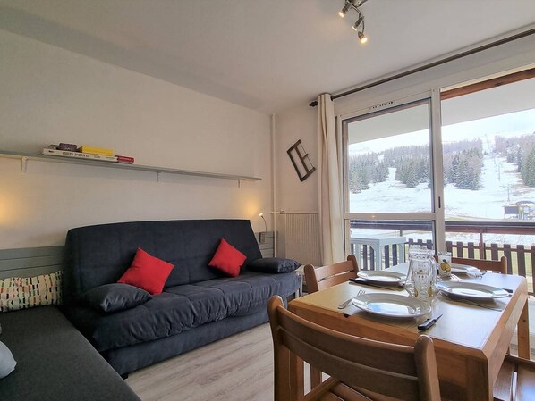 A cozy living room with a dark grey sofa, red cushions, a wooden dining table set for two, and a large window offering a snowy mountain view.