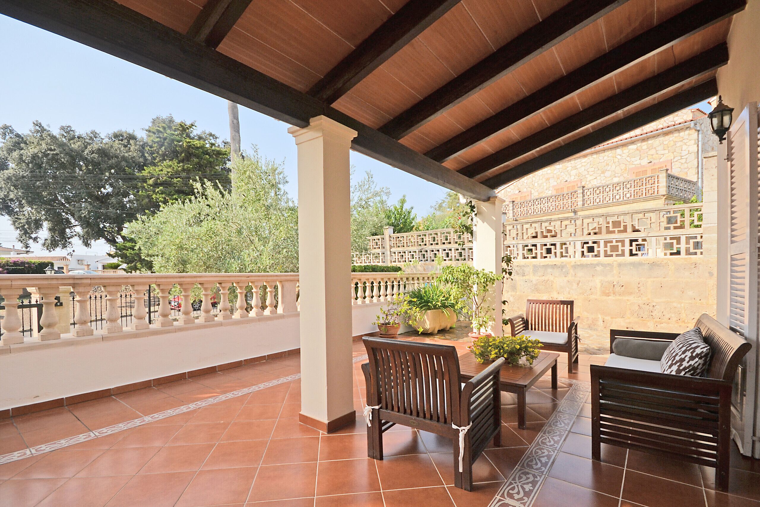 A covered outdoor patio with tiled flooring, wooden furniture, and a decorative railing.