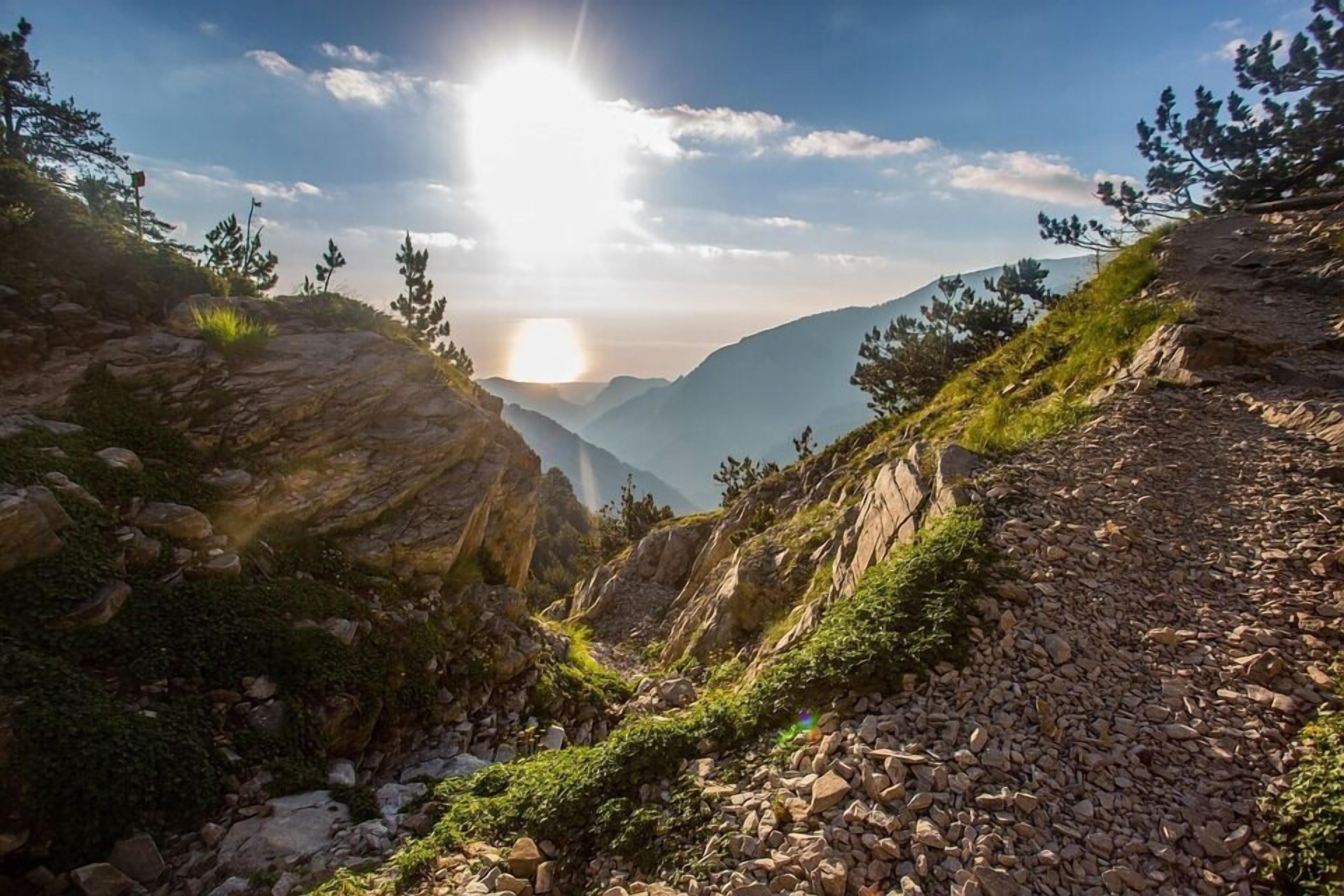 Un sentier de montagne rocailleux offrant une vue sur des montagnes lointaines et sur le soleil se couchant ou se levant.