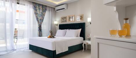 A modern hotel room with a large bed, a nightstand, a desk with a chair, a window with sheer curtains, and a minibar.