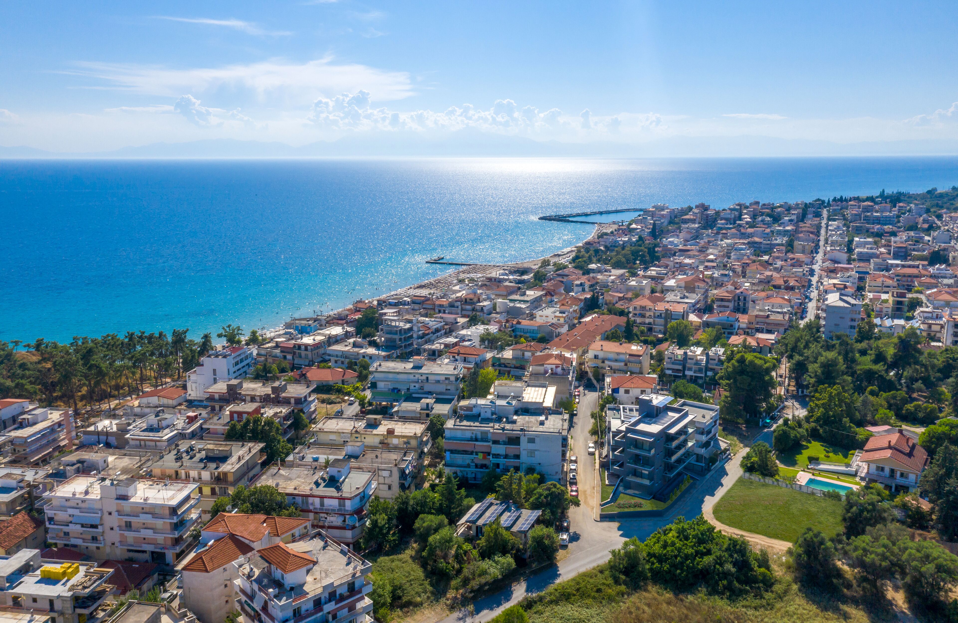 An aerial view of a coastal town with buildings, a clear blue sea, and a distant pier.
