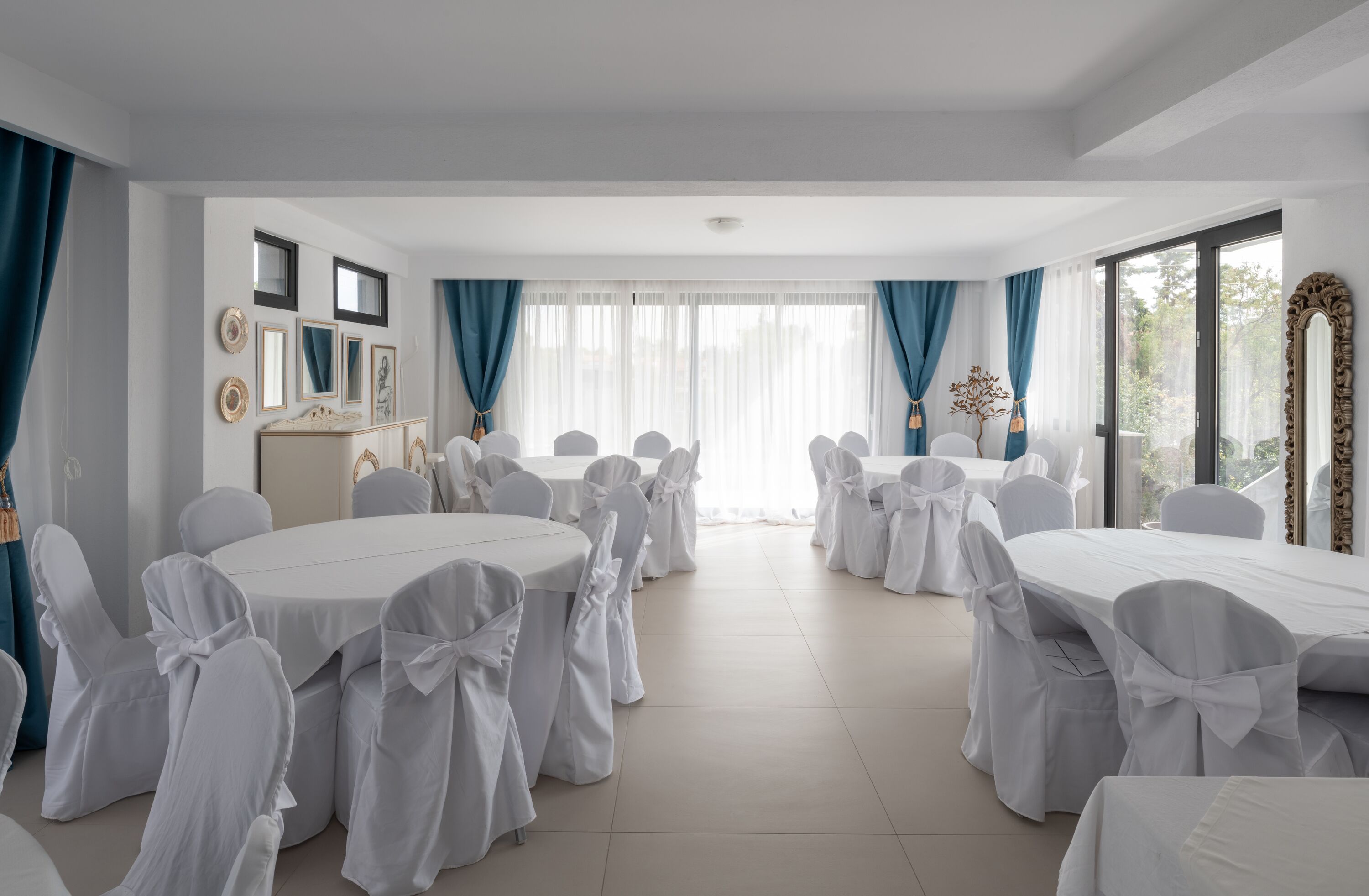 Uma sala de conferências com mesas redondas e cadeiras brancas, grandes janelas com cortinas azuis e um quadro branco na parede esquerda.
