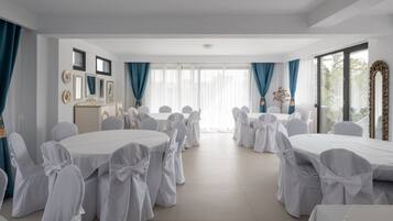 Uma sala de conferências com mesas redondas e cadeiras brancas, grandes janelas com cortinas azuis e um quadro branco na parede esquerda.