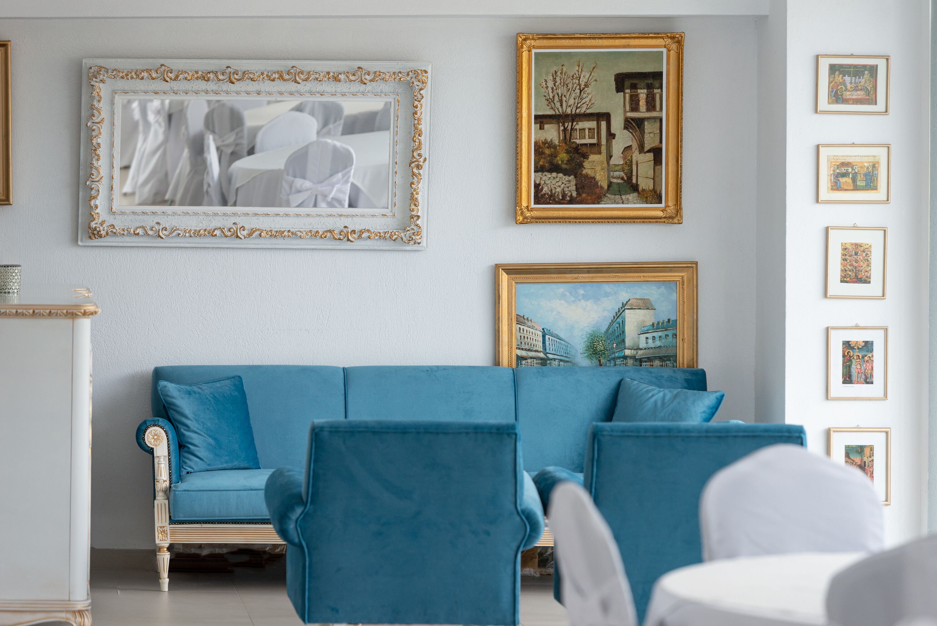 Una habitación con un sofá azul, una silla blanca y una mesa blanca. Hay cuadros enmarcados en la pared.