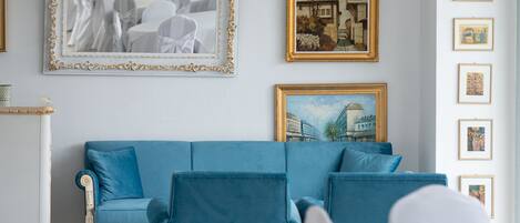 Una habitación con un sofá azul, una silla blanca y una mesa blanca. Hay cuadros enmarcados en la pared.
