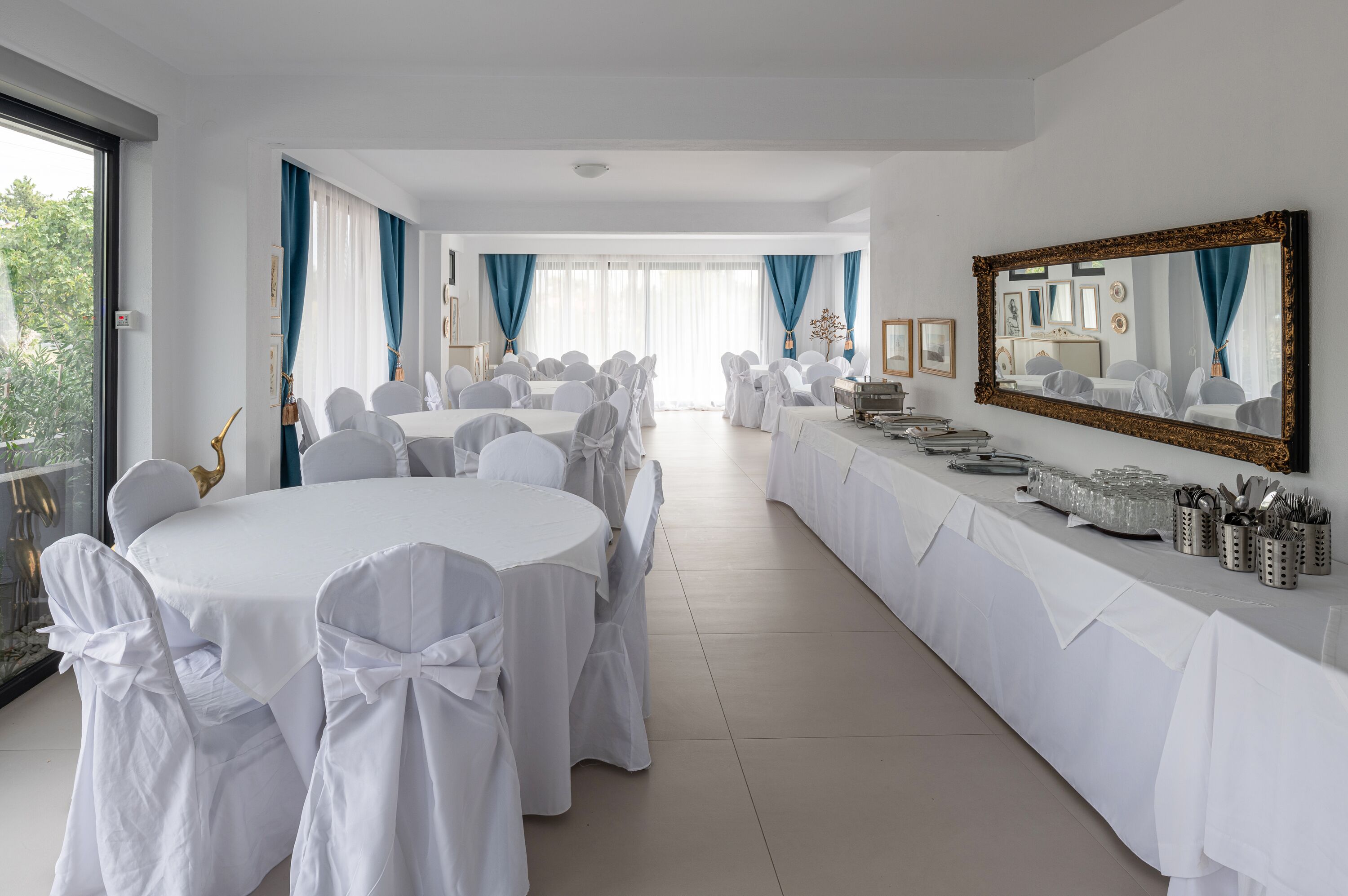 A banquet hall with round tables and white chairs, a long buffet table, and a large mirror on the wall.