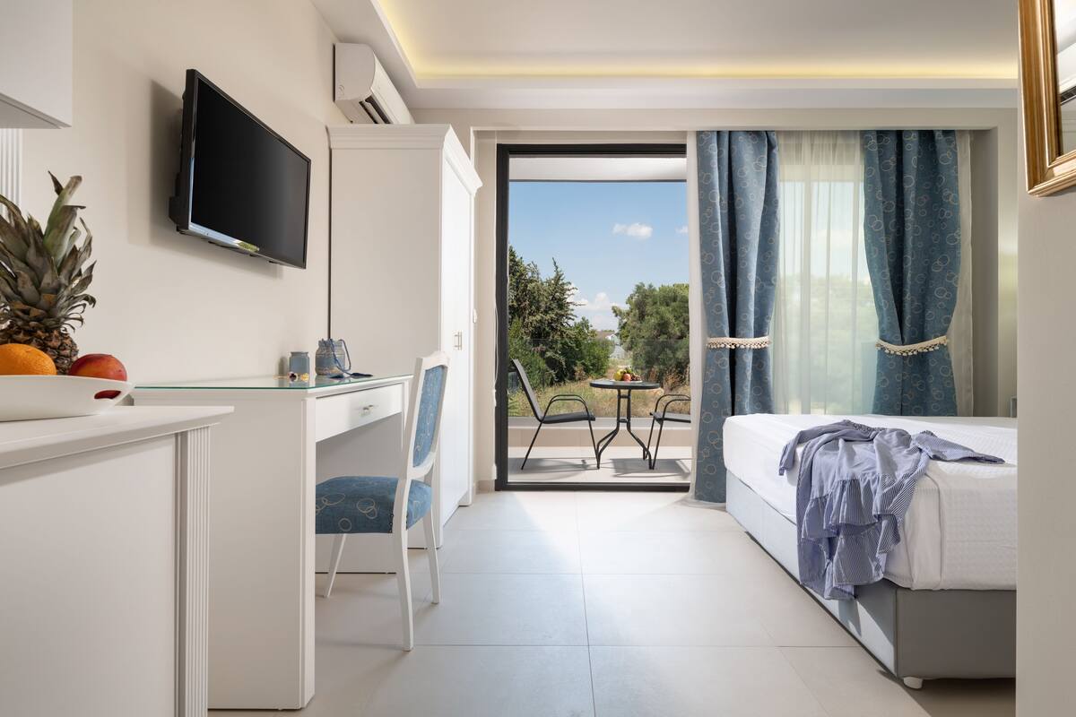 A hotel room with a bed, a desk with a chair, a TV, a balcony with a table and chairs, and a view of the outdoors.