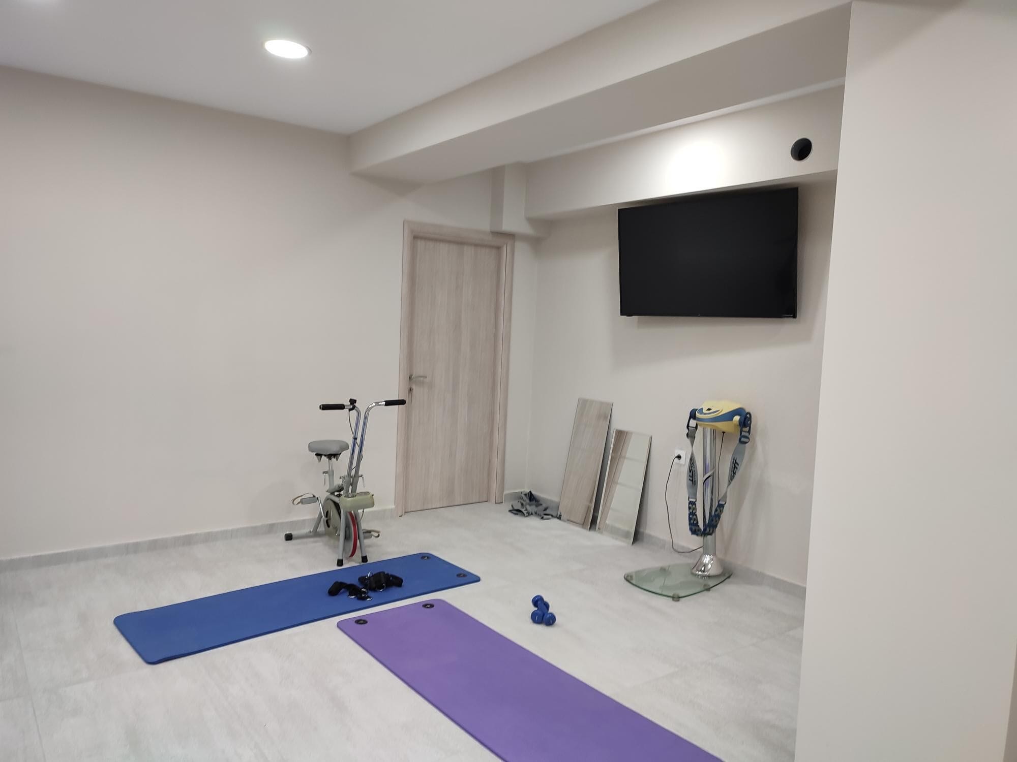 Um quarto espaçoso com ventoinha no teto, televisão na parede e equipamentos de ginástica, incluindo bicicleta ergométrica e tapetes de yoga.
