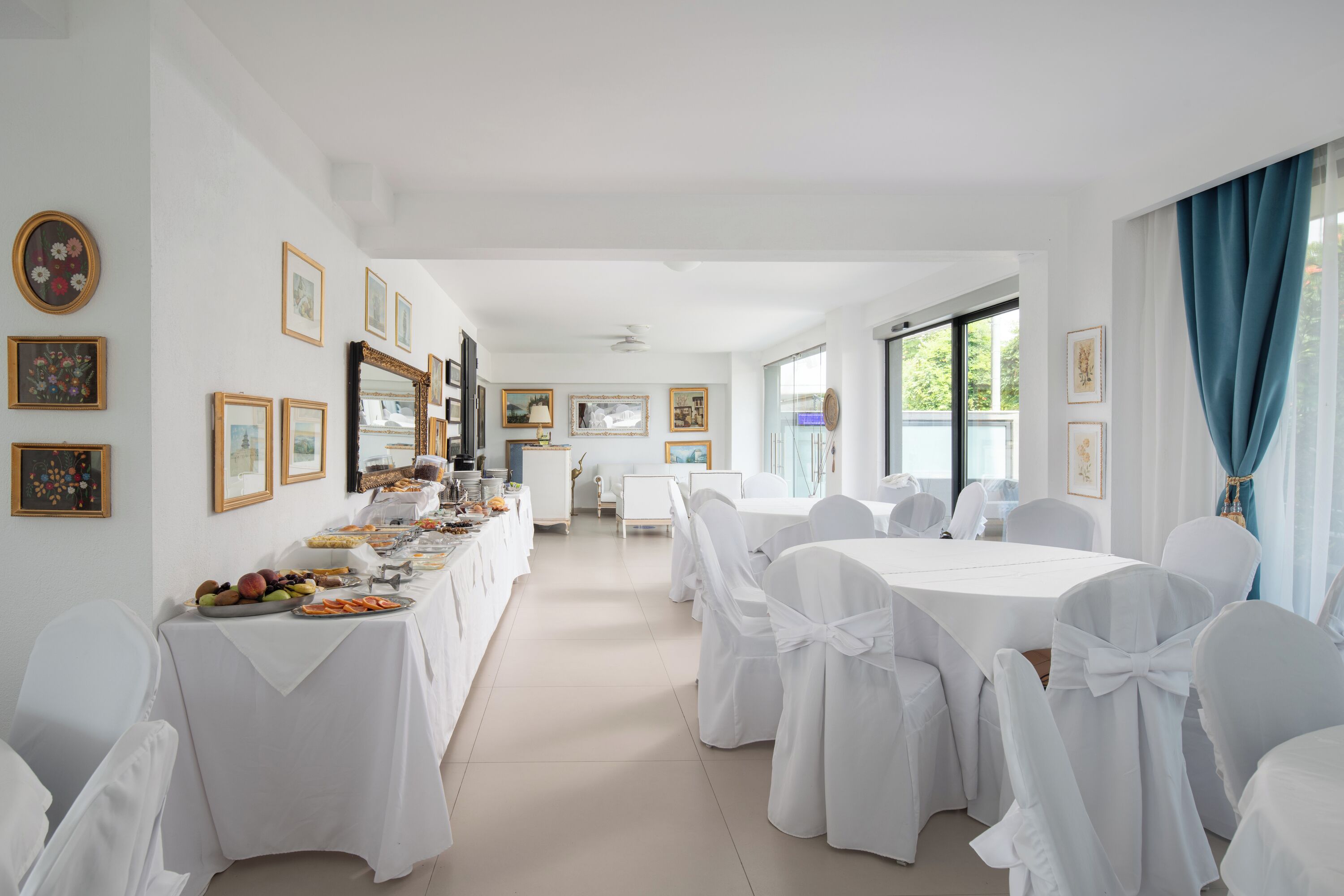 Um quarto amplo com uma mesa comprida posta para refeição, cadeiras brancas e quadros nas paredes.