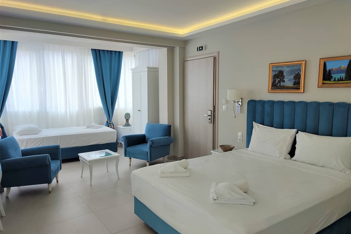 A hotel room with a large bed, two blue armchairs, a small table, and a desk. There is a door with an emergency exit sign, and two framed pictures on the wall.