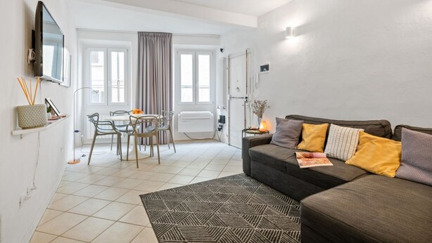 A modern living room with a sofa, a dining table with chairs, a TV mounted on the wall, and a large window with curtains.
