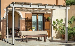 Relax in a serene outdoor seating area under a shaded pergola, surrounded by lush greenery and a building.