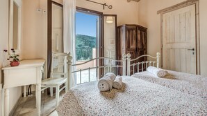 Une chambre avec deux lits, une armoire en bois, un bureau blanc agrémenté d’une plante en pot et une grande fenêtre donnant sur un paysage verdoyant.