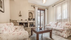 Un salon avec deux canapés à motifs floraux, une table basse en bois, une cheminée en pierre et une vue sur l’extérieur, visible à travers une porte vitrée.