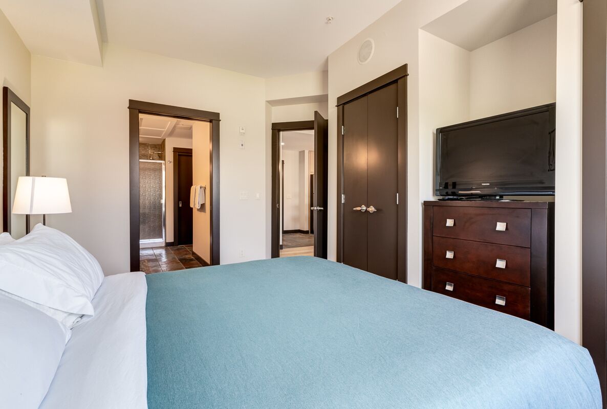 A hotel room with a bed, a dresser, a television, and a door leading to another room.