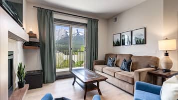 Ein Wohnzimmer mit einer Couch, einem Couchtisch und einem Fernseher. Ein Fenster mit Vorhängen bietet Blick auf Berge.