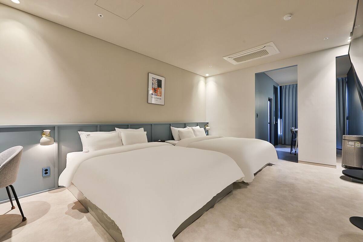 Een moderne hotelkamer met twee grote bedden, een bureau en een stoel. Er hangt een foto aan de muur en er is een deur naar een andere kamer.