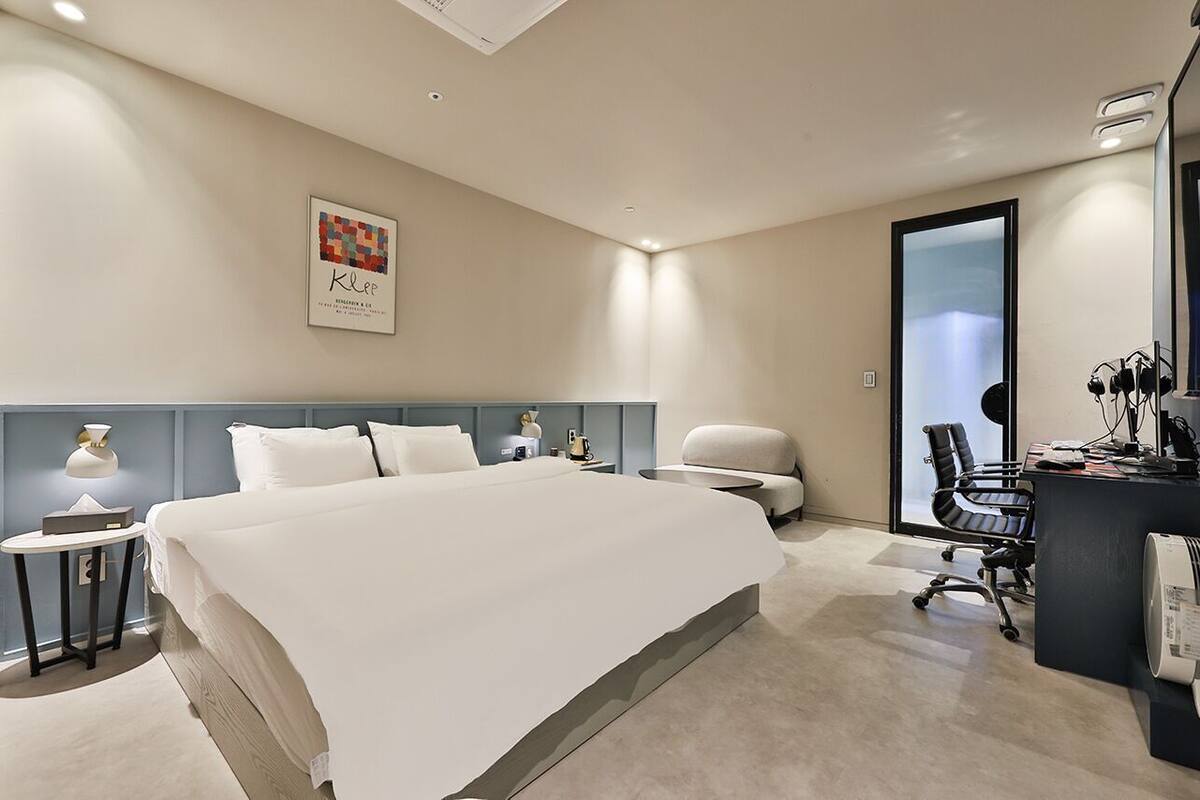 Een moderne hotelkamer met een groot bed, een bureau met computer, een stoel en een kleine tafel met lamp.