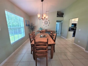 A dining area with a wooden table and chairs, a chandelier, and a window with blinds. - 3BR Marco Island home with heated pool, WiFi, and full kitchen. (Marco Island)