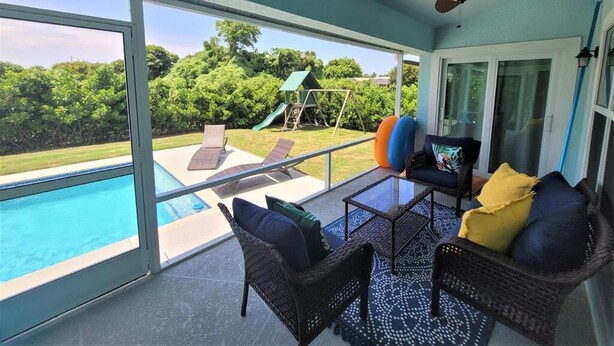 Portico coperto con mobili in vimini, un tavolo in vetro e vista su una piscina e un giardino.