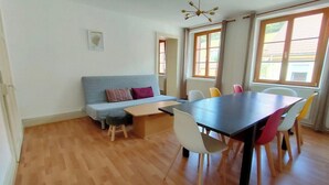 A room with a wooden floor, a grey sofa with a small table, a dark table, and colorful chairs. - Apartment with a view in the center of Bussang (Bussang)