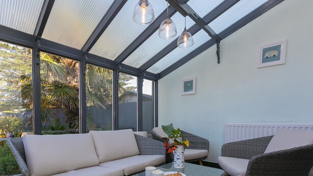 A sunroom with a glass ceiling, a sofa, two armchairs, a glass coffee table, and a vase with flowers.