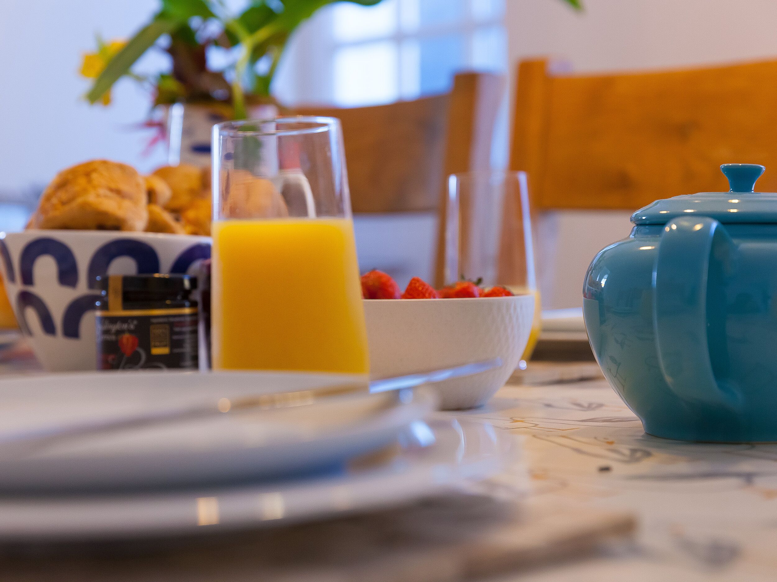 A breakfast setting with a bowl of strawberries, a glass of orange juice, and a blue teapot on a table.