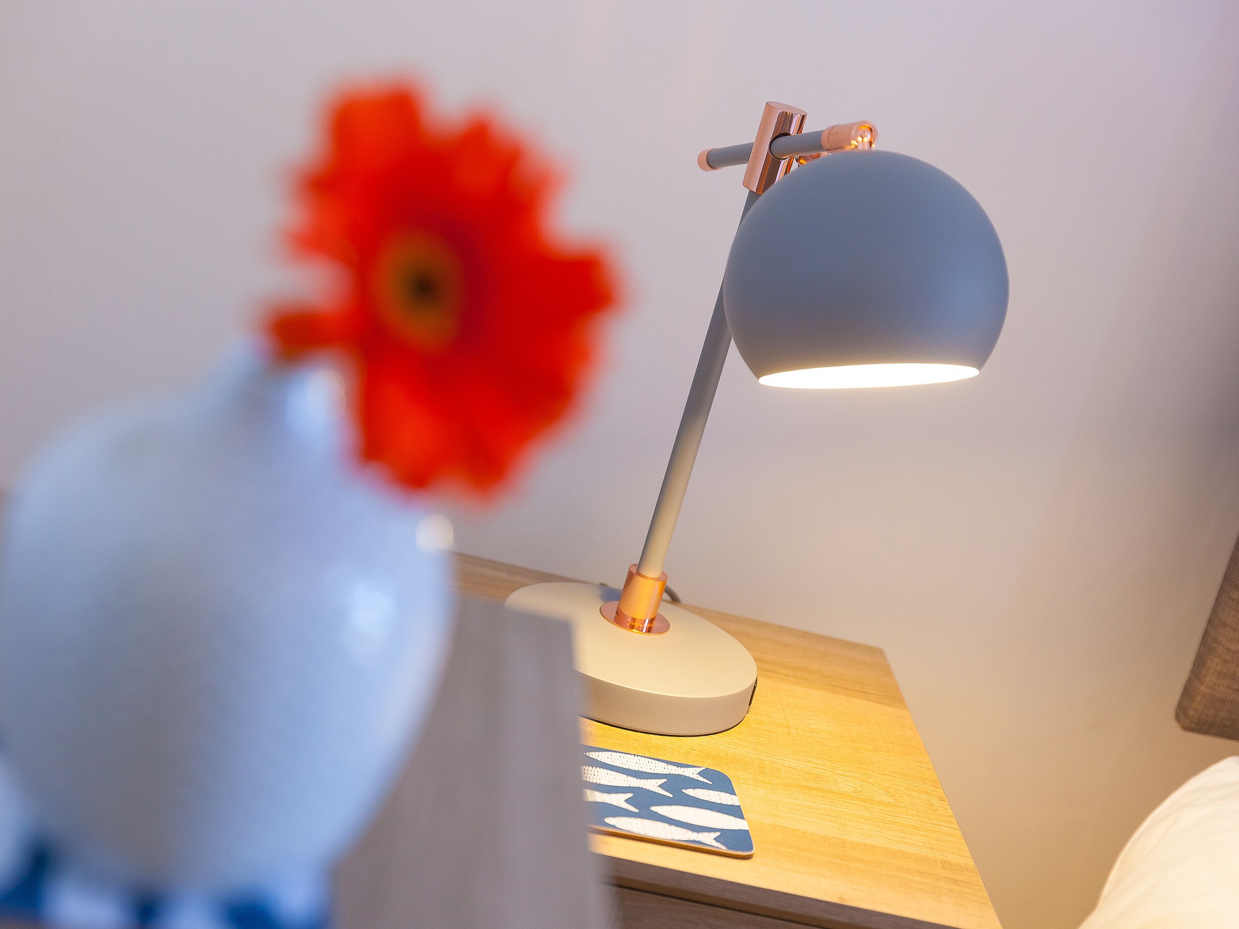 A desk lamp with a white base and a blue shade, a red flower in a vase, and a blue and white patterned coaster on the desk.