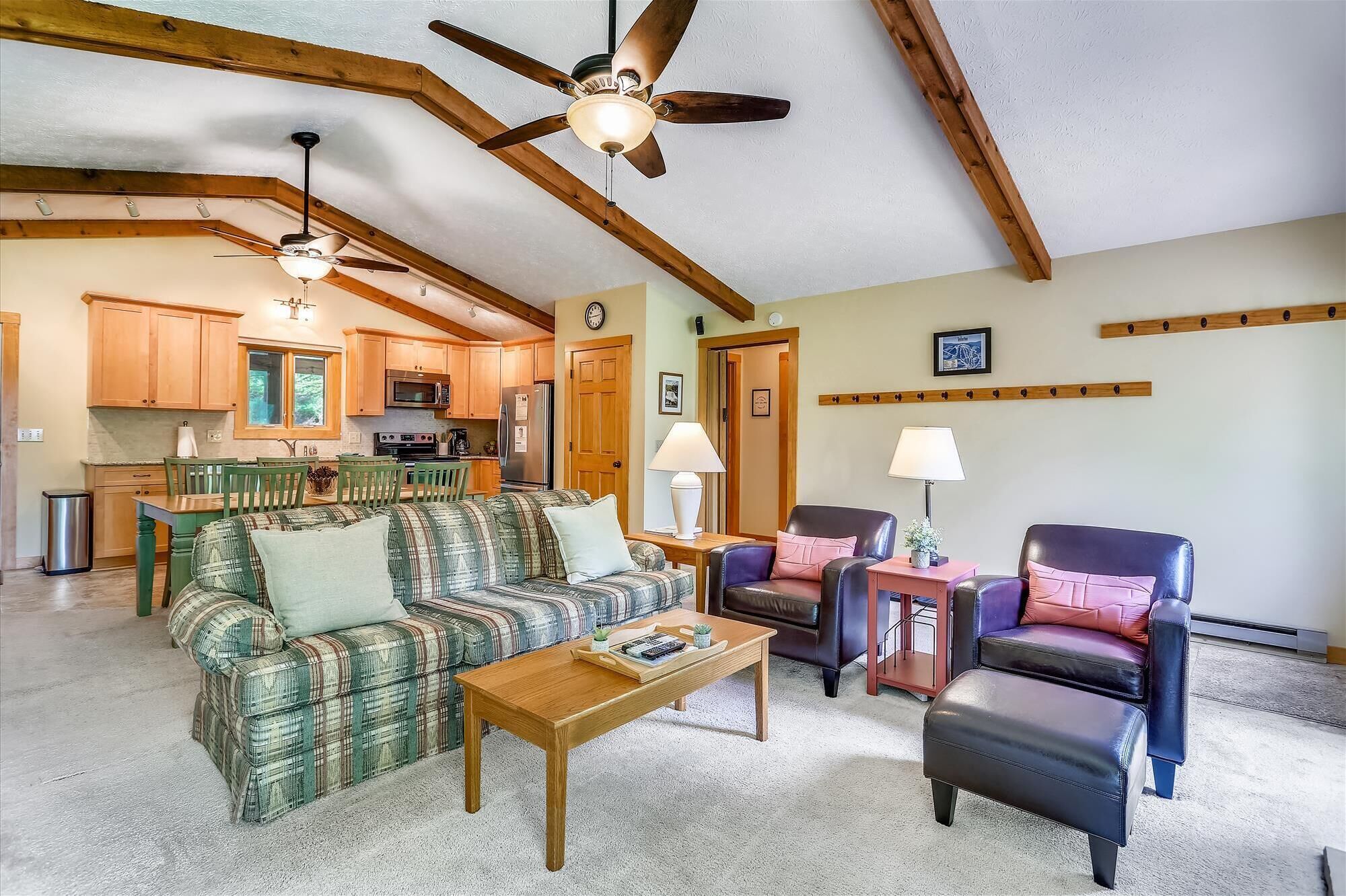 A cozy living room with a plaid sofa, a coffee table, and two chairs. A kitchen with wooden cabinets and a refrigerator is visible in the background.