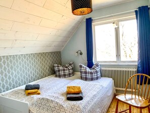 A bedroom with a sloped ceiling, a bed with patterned bedding, two pillows, a chair, a window with blue curtains, and a radiator.