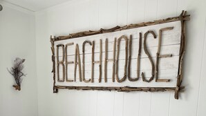Ett sovrum med en säng med mönstrat sänglinne, en väggmonterad skylt med texten ”BEACHHOUSE” och en liten prydnadsväxt.