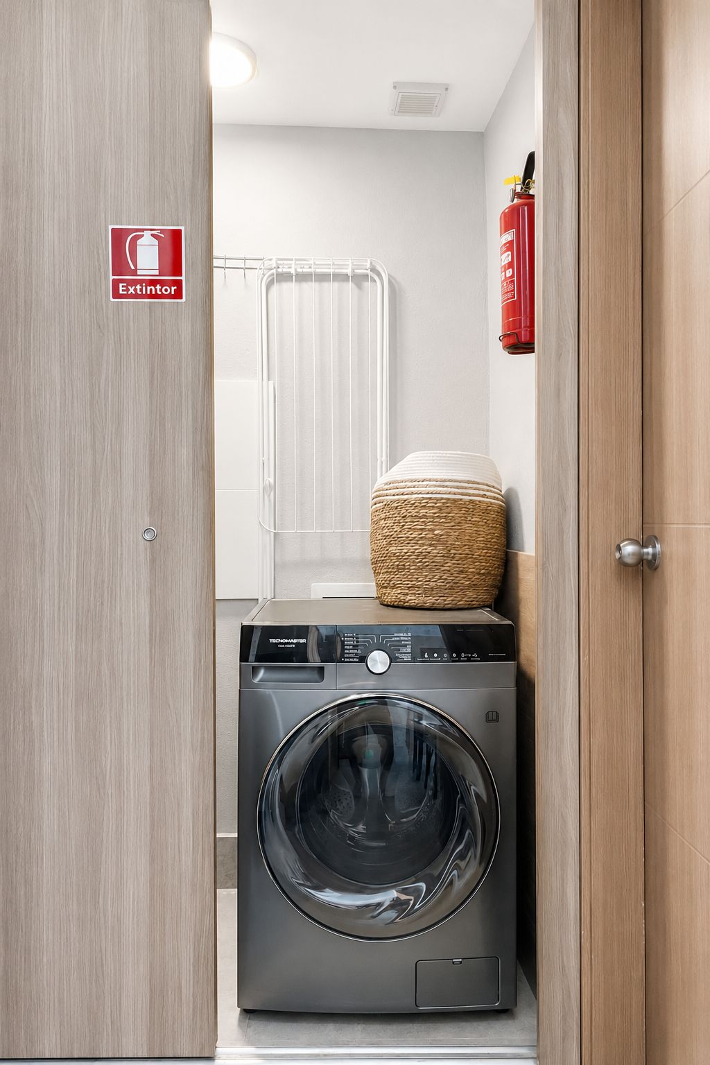 A compact laundry room with a washing machine, a fire extinguisher, and a towel rack.