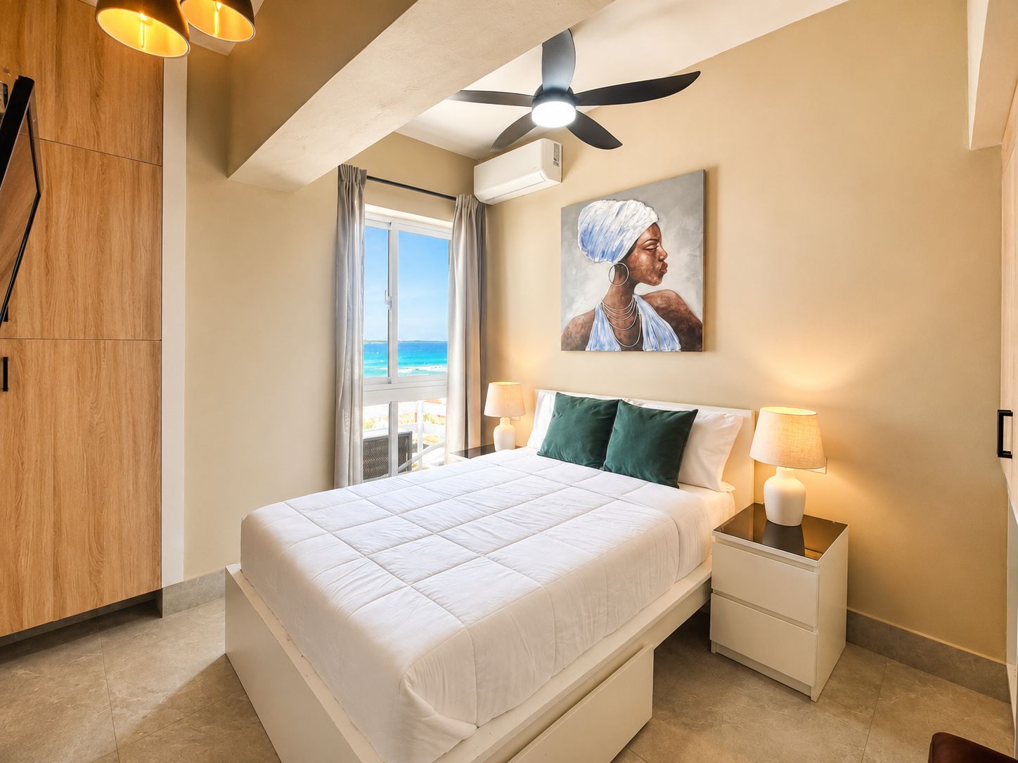 A hotel room with a bed, a ceiling fan, a painting, and a view of the ocean.