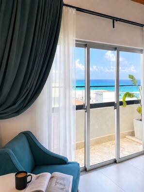 Een kamer met een blauwgroene bank, een koffiekop en een open tijdschrift. Er is een grote schuifglazen deur die leidt naar een balkon met uitzicht op de oceaan.