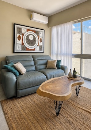 A living room with a grey sofa, a wooden coffee table, and a large window with curtains.