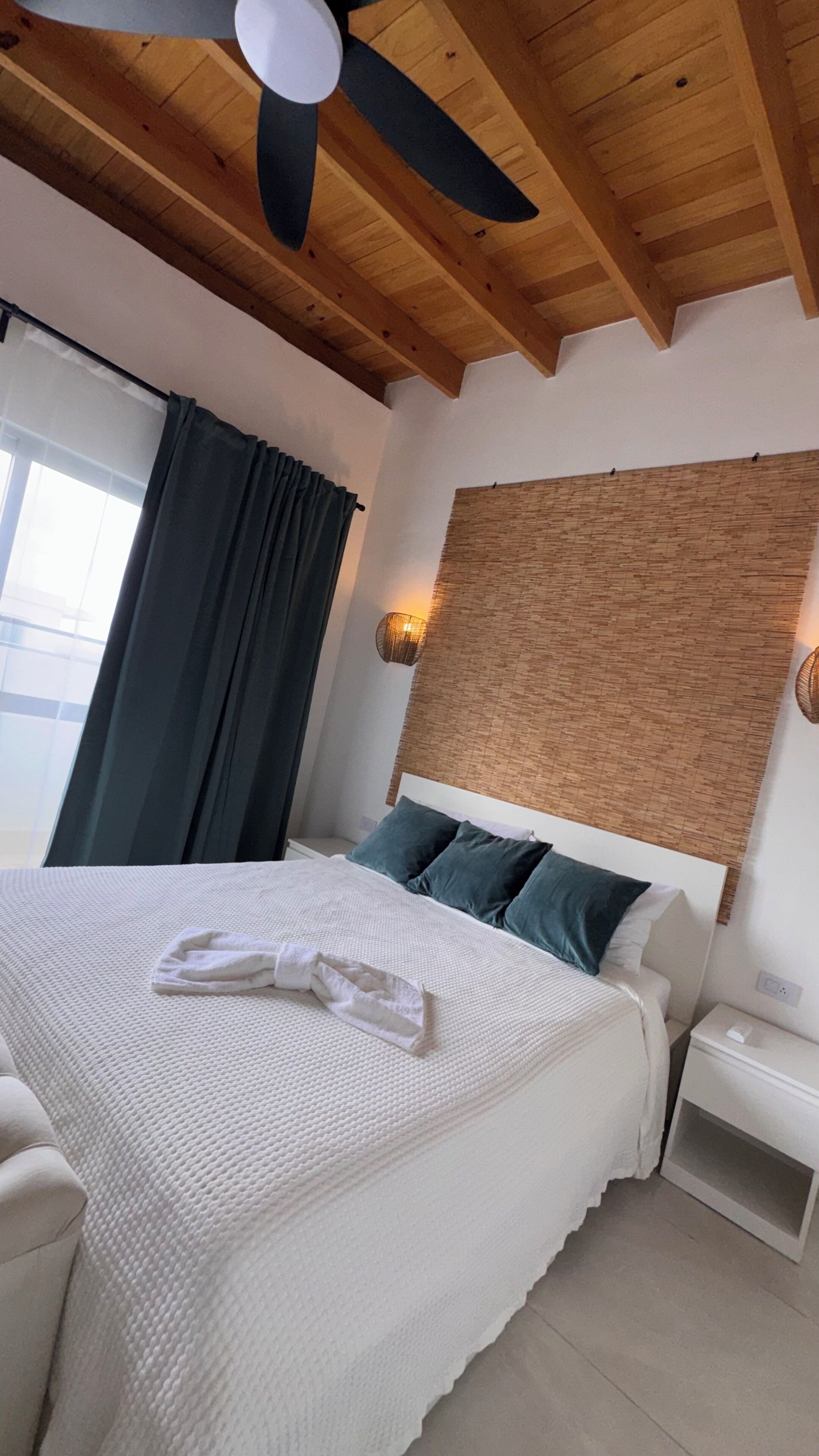 Un dormitorio con cama, ventilador de techo, ventana con cortinas, mesita de noche y lámpara de pared.