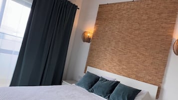 A bedroom with a bed, a ceiling fan, a window with curtains, a bedside table, and a wall-mounted light fixture.