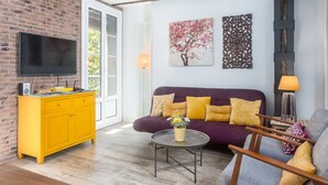 A living room with a brick wall, a flat-screen TV, a yellow cabinet, a purple sofa with yellow cushions, a round coffee table with a vase, and a wooden chair. - Sarriegi | The Rentals Collection (San Sebastián)