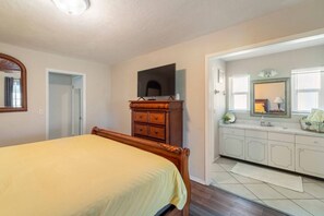 Ein Schlafzimmer mit einem Bett, einem Holzschrank, einem Fernseher, einem Badezimmer mit Waschbecken und Spiegel sowie einer Tür zu einem weiteren Raum.