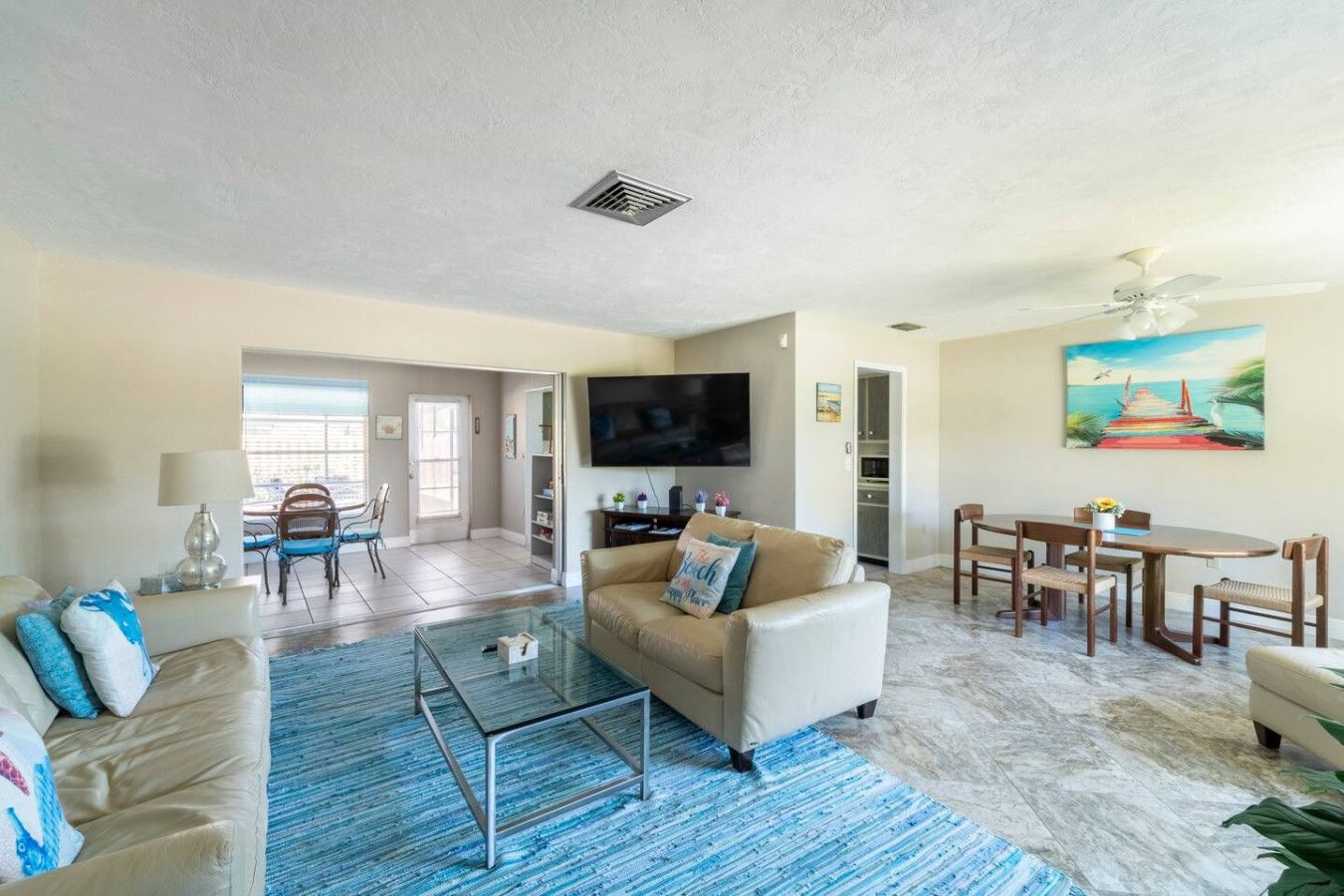 A spacious living room with a beige sofa, a glass coffee table, and a blue patterned rug. There is a dining area with a wooden table and chairs, and a kitchen in the background.