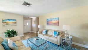 A living room with a beige sofa, a glass coffee table, and two lamps. There are two paintings on the walls and a door leading to another room. - Sarasota Getaway Near Siesta Key, Screened Lanai + Garage, Pet Friendly (Sarasota)