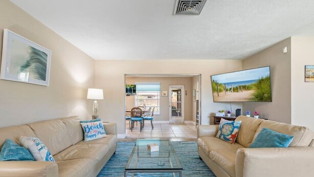 A living room with two beige sofas, a glass coffee table, and a TV on the wall. There is a dining area with a table and chairs in the background. - Sarasota Getaway Near Siesta Key, Screened Lanai + Garage, Pet Friendly (Sarasota)