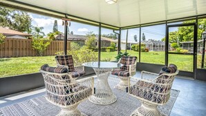 A covered patio with wicker furniture and a glass table, overlooking a backyard with a wooden fence and greenery. - Sarasota Getaway Near Siesta Key, Screened Lanai + Garage, Pet Friendly (Sarasota)
