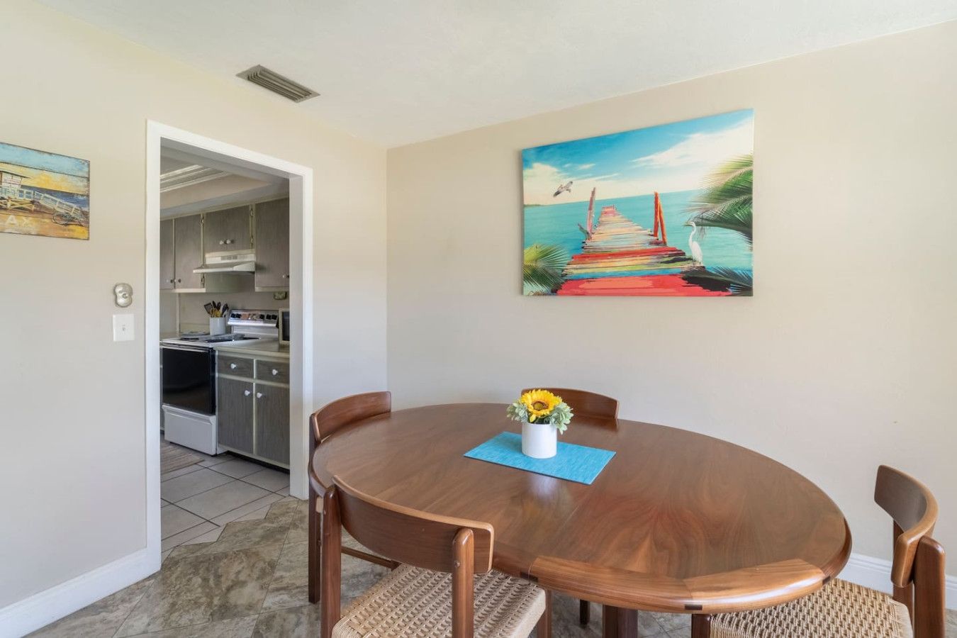 A dining area with a wooden table, four chairs, and a painting of a pier.