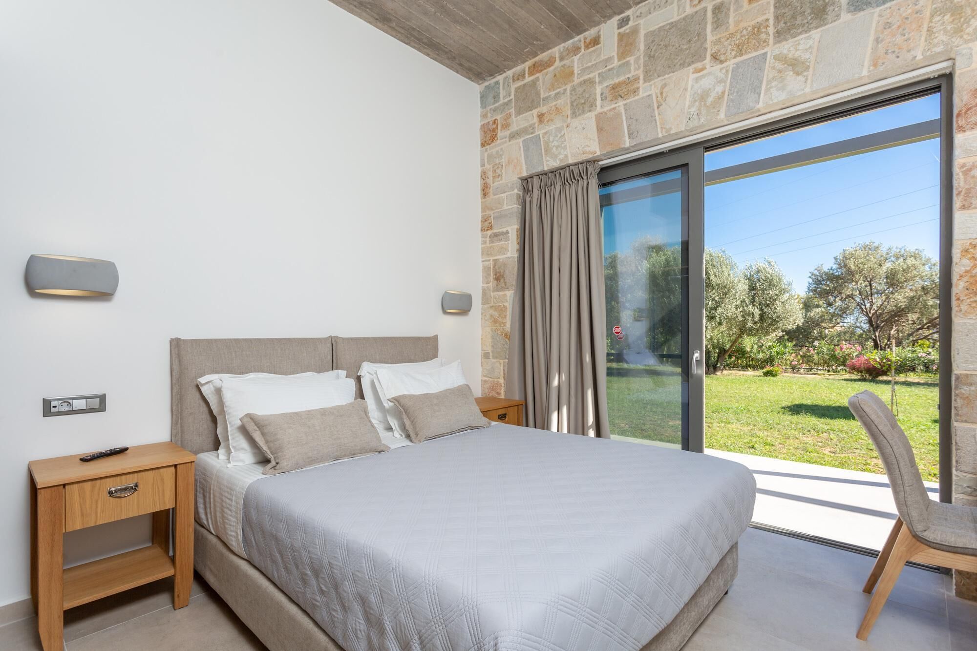 A bedroom with a large bed, a nightstand, a chair, and a view of a garden through a glass door.