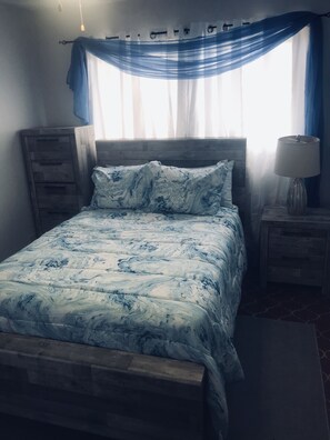 Une chambre à coucher avec un lit, une tête de lit en bois, une table de chevet avec une lampe, une commode et une fenêtre avec des rideaux.